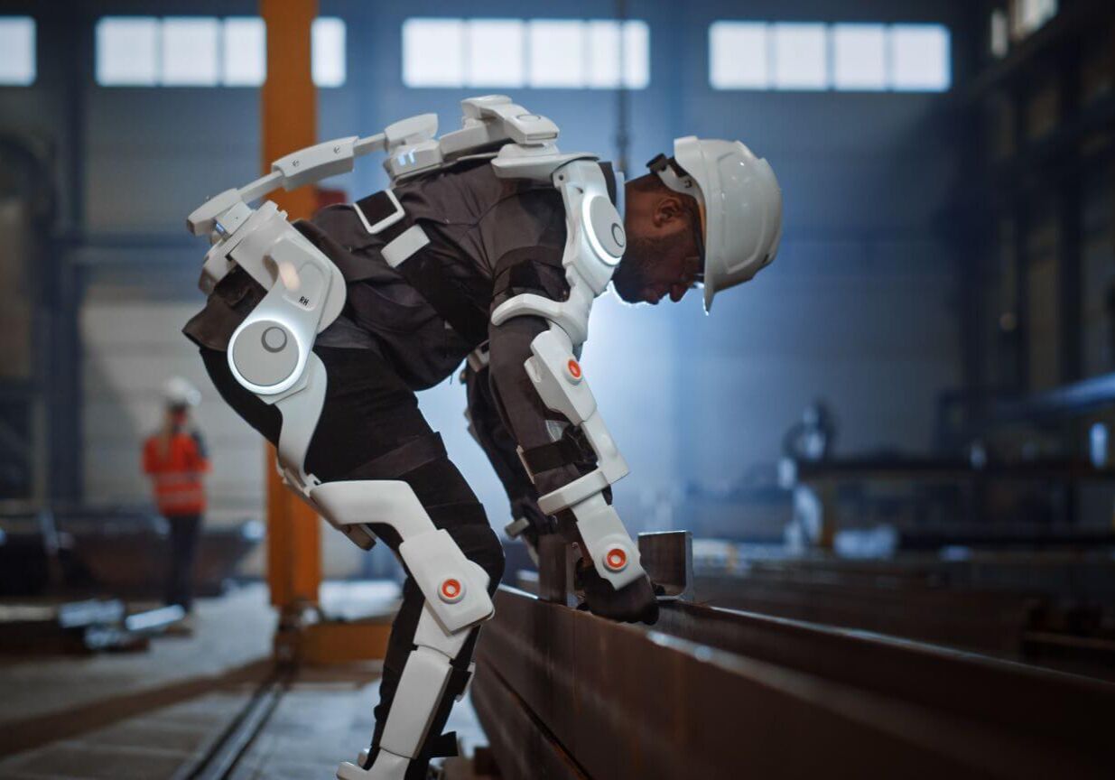 Black African American Engineer is Testing a Futuristic Bionic Exoskeleton and Picking Up Metal Objects in a Heavy Steel Industry Factory. Contractor is Heavy Lifting Steel Parts in a Powered Shell.