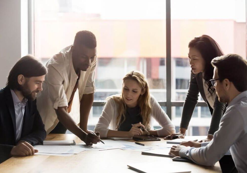 Focused multiracial businesspeople gather at desk brainstorm discuss company financial paperwork at meeting together, concentrated diverse colleagues talk negotiate at briefing in boardroom