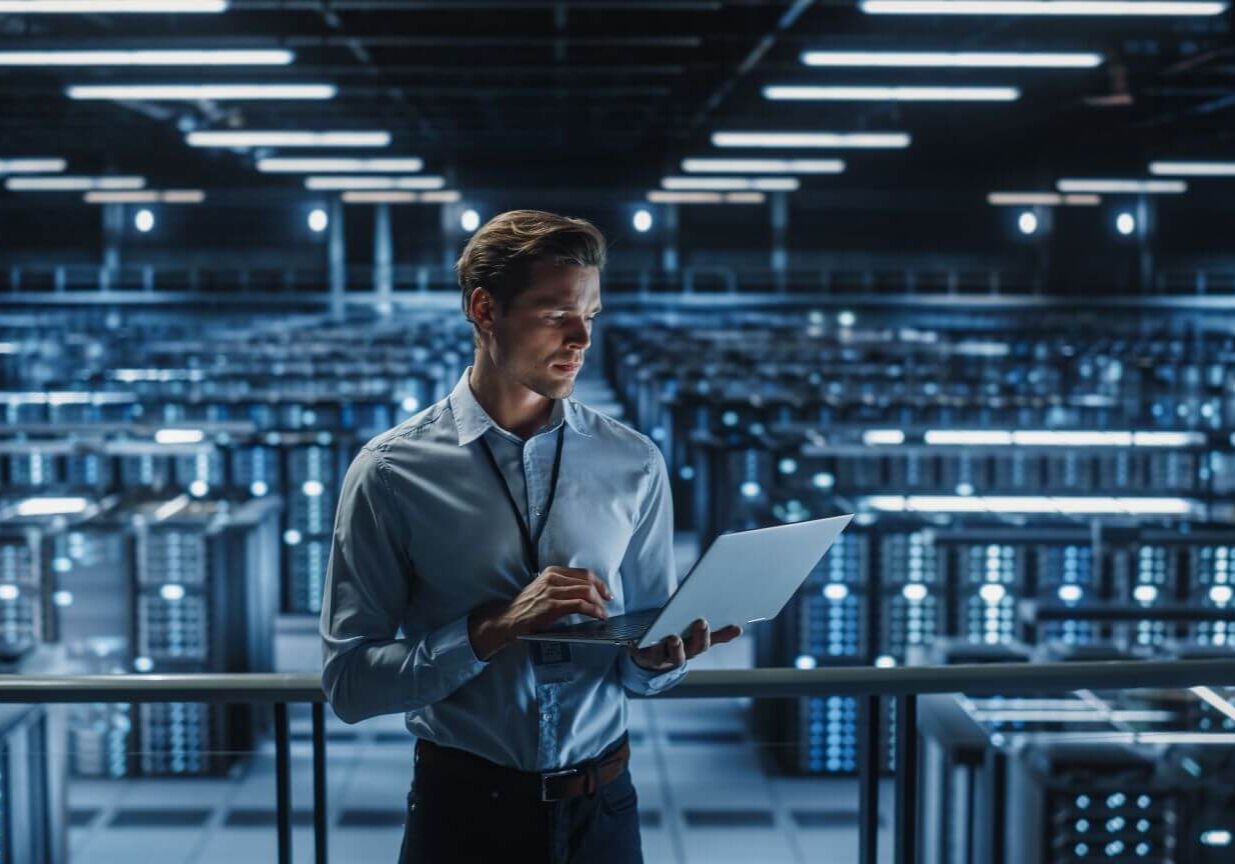 Portrait of IT Specialist Uses Laptop in Data Center. Server Farm Cloud Computing Facility with Male Maintenance Administrator Working on Cyber Security and Network Protection.