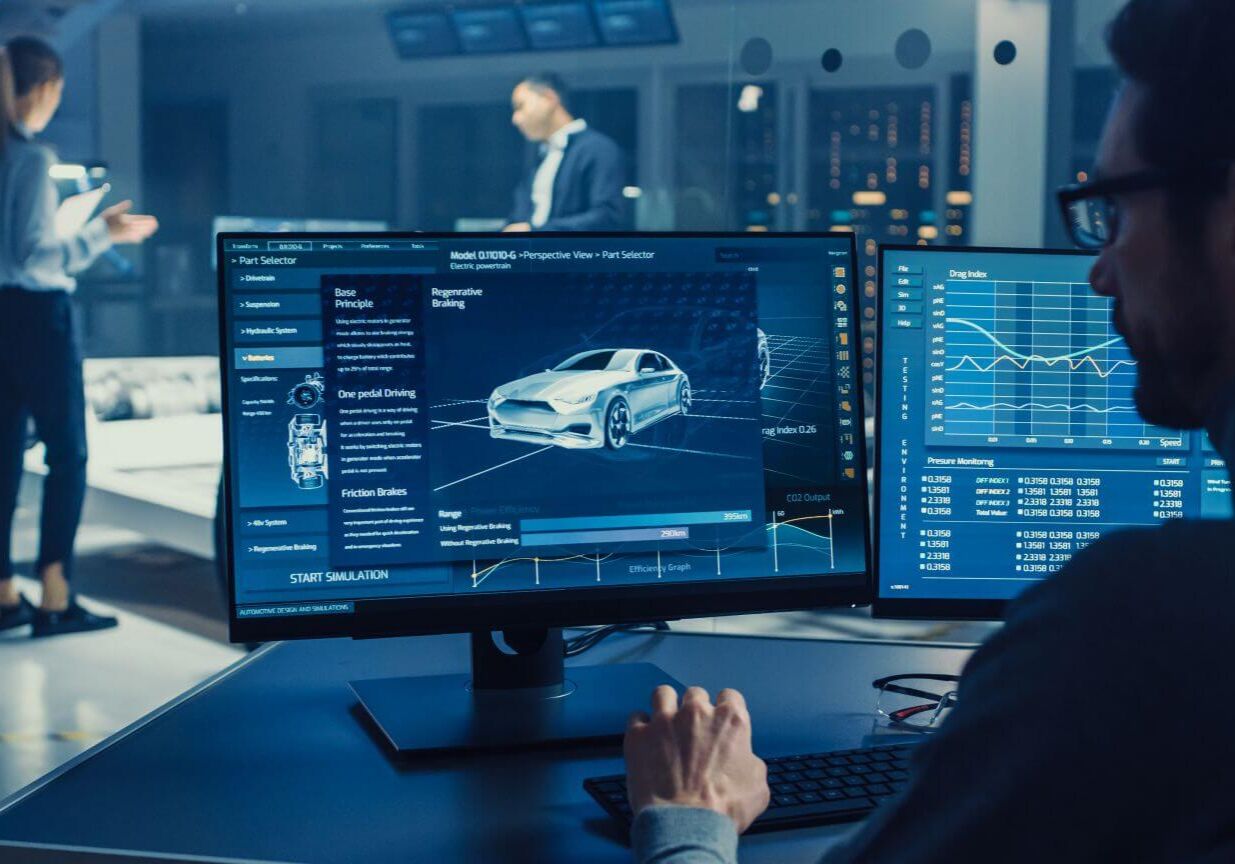 Professional Engineer Works on a Computer with a 3D CAD Software and Tests the Electric Car Chassis Prototype with Wheels, Batteries and Engine Standing in a High Tech Development Laboratory.